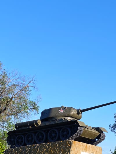 A Soviet T-34 tank is on display, mounted on a pedestal. The tank is painted olive drab and features a white star with a red outline on its turret. The tank's long barrel is pointed to the right. The scene is set against a clear blue sky. To the left, a tree with bare branches and some green foliage is visible. The pedestal appears to be made of rough, light-colored stone. The image was taken in the city of Zimovniki, Russia.