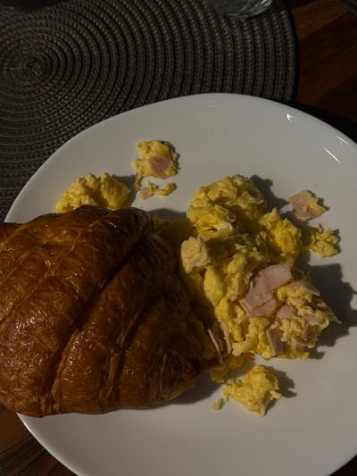 This image shows a close-up, top-down view of a breakfast plate with scrambled eggs and a croissant. The eggs are bright yellow with small pieces of pink ham mixed in. They are piled on the left side of a white plate. A large, golden-brown croissant occupies the right side of the plate, partially obscuring the eggs. The plate is set on a dark, textured placemat, which in turn rests on a wooden surface, likely a table. The lighting suggests it could be morning or evening, with shadows cast by the food indicating a light source from above and slightly to the left. There is no visible text in the image. The scene depicts a simple, comforting meal.