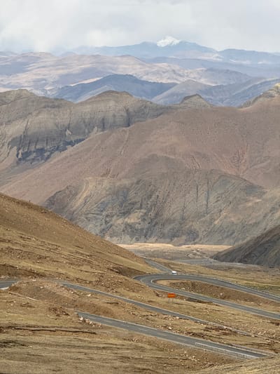 The image depicts a winding road traversing a mountainous landscape in what appears to be Zagor town, China. The road features a series of hairpin turns, ascending the steep incline. The terrain consists of barren mountains with varying textures and geological features. The background showcases a range of distant mountain peaks shrouded in haze. A few vehicles, including a white truck, are visible on the road. A red sign is placed on the road.