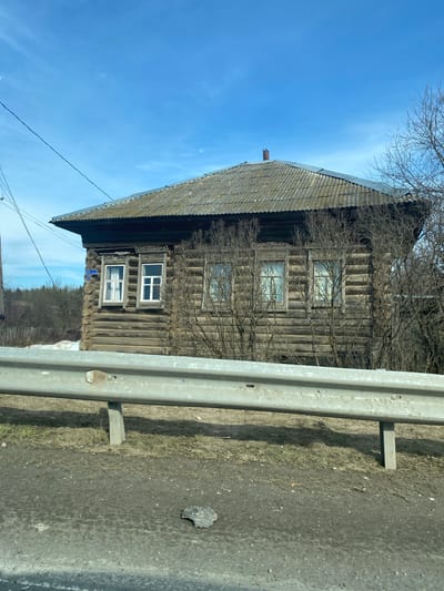 A two-story log house stands next to a roadside barrier in Malyi Dubovik, Russia. The house has a steeply pitched, dark gray roof. Three window sets are visible on the house's front facade. Bare trees stand near the house. Blue sky with a few clouds fills the top left of the frame. A gray metal roadside barrier stands prominently on the right. The road surface is dark and slightly damp. Power lines are visible against the sky.