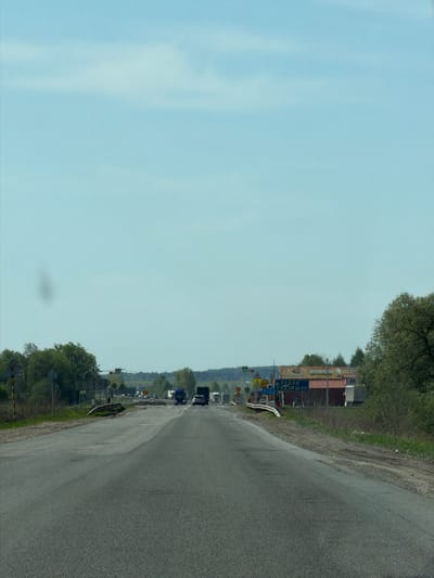 The image shows a road in Совхозный, Russia, with traffic and signage along the roadside. The road surface is asphalt and appears to be slightly weathered. There are several vehicles visible on the road ahead, including what appears to be at least one truck and several cars. A dense collection of road signs and advertising boards are lined up on the right side of the road with Russian lettering. The sky is a light blue and there are green trees and bushes on either side of the road.