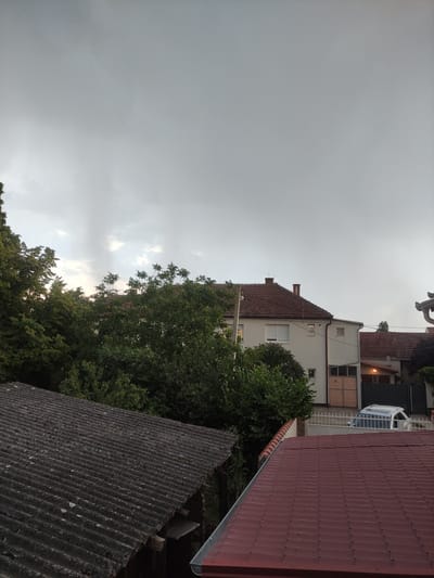 The image depicts an overcast sky above a residential area in Zrenjanin, Serbia. The perspective is from a rooftop, looking across several buildings and dense green foliage. In the foreground, a dark, corrugated roof is visible, possibly made of asbestos cement, showing signs of age and weathering. Beyond this, a red-tiled roof of a modern house extends into the frame.
The middle ground features buildings with various architectural styles. One building is primarily white with a red-tiled roof and prominent chimneys. It has several windows, some with white shutters. Adjacent to this, another structure appears to have a dark facade and a window with interior lighting suggesting it might be evening. A greenhouse-like structure with a curved roof and metal supports is also visible. Trees with lush green leaves dominate the space between the buildings, their branches reaching towards the sky.
The sky is uniformly grey, indicating cloudy weather, likely before or during rainfall. The overall atmosphere is somber due to the lack of direct sunlight and the muted colors. The image seems to be taken during the daytime, though the heavy cloud cover obscures any direct indication of the sun's position. No people are visible in the frame. The scene captures a typical urban or suburban landscape under a heavy sky.