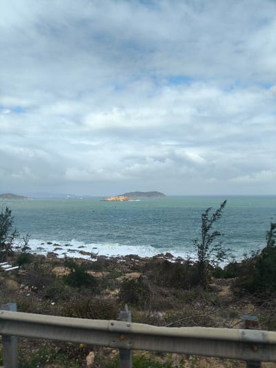 The image depicts an overcast coastal landscape in Tuy Phong 4, Vietnam. In the immediate foreground, a metallic grey guardrail with visible bolts and yellow reflective dots runs horizontally across the bottom. Behind the guardrail, the terrain consists of dry, rocky ground with sparse, low-lying scrub brush, dark green vegetation, and some taller, spindly bushes. White debris is visible on the rocky ground near the water's edge on the left.
The midground features a body of teal-green ocean water with moderate waves breaking into white foam against a rocky shore. In the middle distance, several landmasses are visible. A prominent, rocky island with patches of brown exposed rock and dark green vegetation is situated to the right. Further left, a larger, more elongated landmass covered in dark green vegetation is present. Small, indistinct white vessels or buoys are scattered on the water between these landmasses and the foreground. The sky above is predominantly covered with a thick layer of grey and white stratocumulus clouds, with small, scattered patches of visible blue sky. No individuals or discernible actions are present.