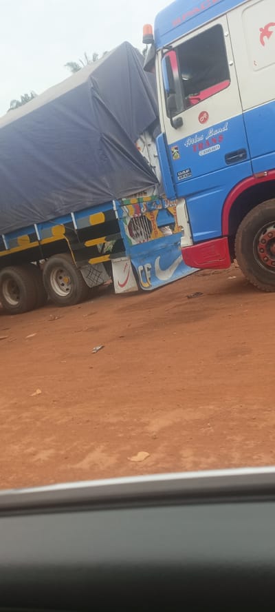 The image shows a blue and white DAF XF 430 truck with the name "Atlas Land Trans" painted on the cab. The truck is partially covered by a blue tarp, likely transporting goods. Its flatbed sides are decorated with a white Nike swoosh. The ground is reddish-brown dirt, suggesting an unpaved road. The photo was taken in Oto, Nigeria as indicated by a roadside sign. The lower portion of the image is blurred, suggesting it was taken from inside a vehicle.