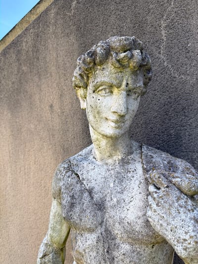 A weathered stone statue of a male figure lies on its side on a flat, concrete surface. The statue depicts a man with curly hair and detailed facial features. The statue is covered in grey and white discoloration, likely from exposure to the elements. The concrete surface is also textured. The photograph was taken outdoors, likely at Oullins-Pierre-Bénite, France.