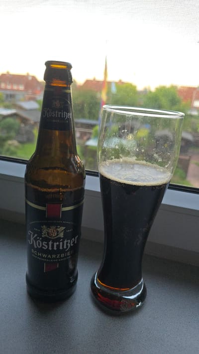 A bottle and a glass of dark beer, likely Köstritzer Schwarzbier, are on a grey surface near a window. The glass is filled with beer, showing a frothy head. The bottle is partially empty and lying on its side. Through the window, a blurred background of houses and greenery can be seen.