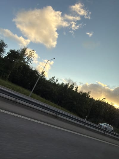 The image, taken from a moving vehicle on a highway, depicts a rural scene near Ödsmål, Sweden. The sky is a pale blue with large, fluffy, white clouds illuminated by the setting sun, which casts a warm golden glow on the clouds on the right side of the frame. A dense line of dark green trees borders the right side of the highway, with the sun's rays piercing through them. A single white car is visible on the right lane of the highway, traveling in the same direction as the photographer. On the left side of the highway, a silver metal guardrail separates the lanes. A tall, grey lamppost stands near the trees on the left side of the road. The highway itself is paved and grey, with white lane markings. The ground beside the highway is covered in green grass and scattered trees, suggesting a natural, undeveloped landscape. The image has a slight tilt to the right, characteristic of being taken from a moving vehicle.
