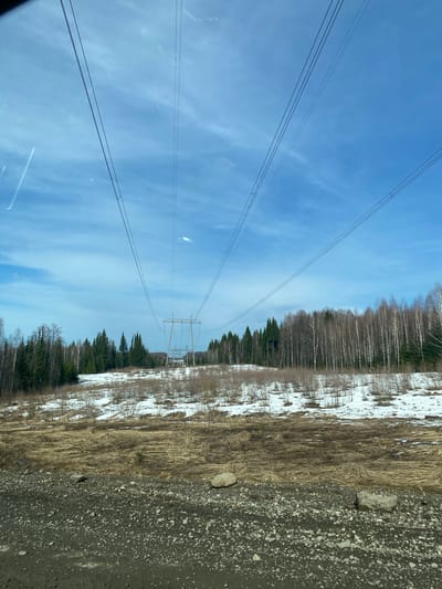 The image shows a rural landscape in Malyi Dubovik, Russia. Overhead power lines stretch across a bright blue sky with scattered clouds. The ground is divided between a gravel road and a grassy area with patches of melting snow. A thin forest of birch trees and coniferous trees runs parallel to the road. An electrical transmission tower is visible in the distance. There are no people or animals visible in the frame.