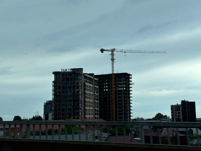The image captures a cloudy day in Makhinjauri, Georgia, focusing on the construction of several buildings. In the foreground, a metal railing runs horizontally across the frame, partially obscuring a view of residential roofs with red tiles. Behind this railing, two large, unfinished apartment buildings dominate the scene. The building on the left has a sign with blue letters that reads "TEKTO". A construction crane, painted yellow and orange, stands tall next to the right building, extending horizontally across the sky. Further in the background, another, more completed dark building is visible, and a line of trees separates the urban landscape from the horizon. The overall impression is one of ongoing development in a coastal or near-coastal urban environment.