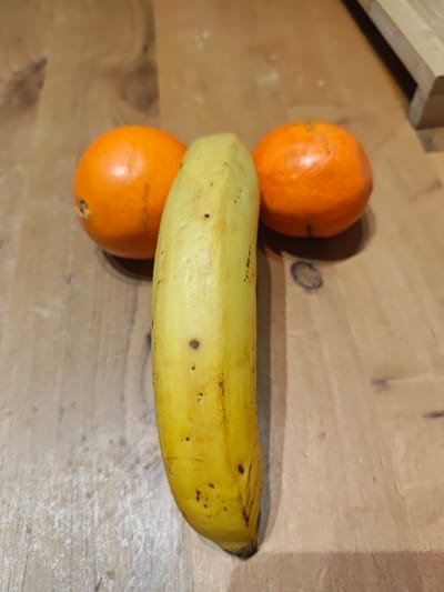 The image displays a still-life arrangement of three pieces of fruit on a wooden surface. Dominating the center of the frame is a single ripe banana, positioned vertically, extending from near the top to the bottom edge of the image. The banana is yellow with several small brown spots and some minor scuff marks on its peel, indicating a natural ripening process.

Flanking the banana, one on each side, are two round, bright orange fruits, which appear to be mandarins or clementines due to their size and characteristic peel texture. The orange on the left is positioned slightly above the mid-section of the banana, while the orange on the right is similarly placed on the opposite side. This particular arrangement of the fruits creates a suggestive, phallic shape.

The background consists of a light brown wooden surface, likely a table or countertop, showing visible wood grain patterns and some subtle variations in color and texture, including what might be faint scuff marks or dust. The lighting appears to be even and consistent with an indoor setting. There is no discernible text or specific time of day indicated. The overall impression is a simple, humorous composition of everyday fruit.