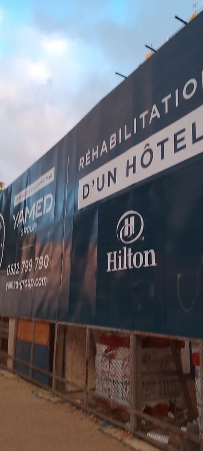 The image displays a large construction banner or hoarding at an outdoor site, indicating an ongoing rehabilitation project. The setting is likely a construction area, with a fence or scaffolding visible below the banner. The sky above is overcast, with shades of light blue and white clouds, suggesting it could be late afternoon or early evening.
The primary subject is the dark blue banner, angled across the frame. Prominently displayed in white capital letters are the words: "RÉHABILITATION D'UN HÔTEL" (Rehabilitation of a hotel). Below this, the distinctive Hilton logo and the "Hilton" brand name are clearly visible, indicating the project is for a Hilton property.
To the left side of the banner, smaller white text reads "PROJET DÉVELOPPÉ PAR YAMED GROUP", identifying the developer. Contact information includes a phone number "0522 799 790" and a website "yamed-group.com". The "0522" prefix is typical for landlines in Casablanca, Morocco, aligning with the provided location context of Aïn El Aouda, Morocco.
Below the banner, a metal mesh fence or frame, likely part of the construction barrier, partially obscures views of the ground and materials behind it. Some indistinct graffiti or markings can be seen on elements behind the fence. The ground in the foreground appears to be unpaved, consisting of dirt or gravel. No people are present in the image.