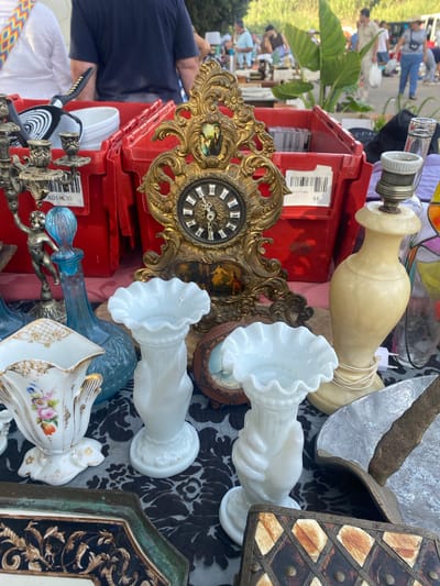 This is a photograph of an outdoor flea market scene in San Luis de Sabinillas, Spain. The foreground is filled with various antique and decorative items displayed on a black and white patterned tablecloth. Prominently featured are two white milk glass vases with ruffled rims, a white porcelain vase with floral decoration, and an ornate brass clock with roman numerals on its face. To the right, a cream-colored lamp with a coiled cord is visible. In the background, red plastic bins are stacked, containing various items including what appears to be a golf club. Several people are casually browsing the stalls in the distance. The setting suggests a bright, sunny day with trees and foliage visible in the background.