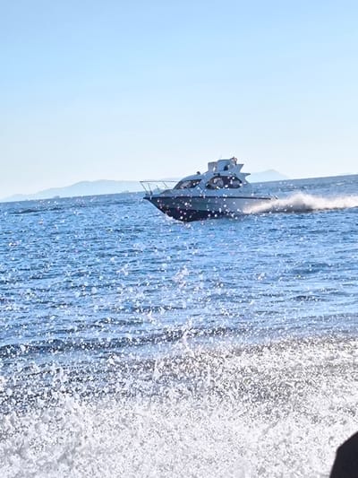 A white motorboat is speeding across a blue body of water, leaving a wake of white spray. The water is choppy, with small waves. In the foreground, there is a large splash of white water. The sky is a clear blue. In the distance, there are hazy mountains. The image is taken from a low angle, suggesting the viewer is on another boat or in the water. There are no people visible on the boat, but there appears to be a dark silhouette of a person in the lower right corner of the frame, possibly the photographer. The scene is set in Pasir Panjang, Indonesia.