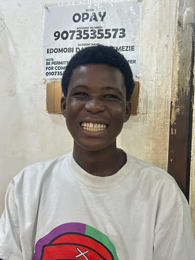A medium shot depicts a young man smiling at the camera. He is wearing a white t-shirt with a graphic design on the right side. Behind him, on the wall, is a poster with the word "OPAY" in large letters, followed by an account number, "9073535573," and the account name, "EDOMOBI DA MEZIE." The poster includes a note with a phone number. The location is inferred to be in Ogwashi-Uku, Nigeria, based on the context of the provided OCR information.