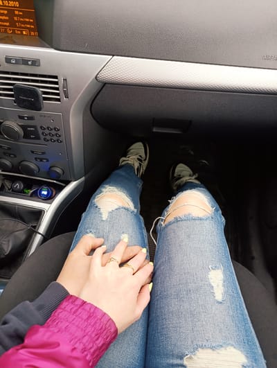 The image shows the interior of a car, specifically the front passenger area. A person is seated, wearing ripped blue jeans and black and white sneakers. Another person is holding their hand. The dashboard is visible, with various car controls, including an older-style CD player and the car's display screen, which shows a date of 8.10.2010. The gear shift is visible, as is a phone charger plugged into the center console. The car make and model are not discernible from the image. There is no evidence of location in Fryanovo, Russia in this image.