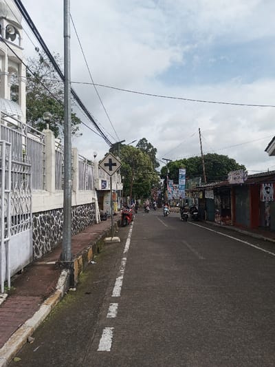 A straight road stretches into the distance with motorbikes traveling along it. A crosswalk sign stands on the left side of the road, next to a building with a fence. Power lines crisscross overhead. Buildings and trees line both sides of the road. The sky is cloudy.