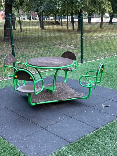 An outdoor merry-go-round with attached seats and a table is situated on a black rubberized playground surface. The merry-go-round features green metal tubing forming the structure and armrests, with brown wooden seats and a brown wooden tabletop. The tabletop has a decorative pattern. Four seats are attached, each with a small yellow accent on the backrest. The merry-go-round is surrounded by artificial turf on the right and natural grass behind it, separated by a green metal fence. In the background, through the fence, are trees and a dirt embankment. The scene appears to be located in a park or playground area in Piatykhatky, Ukraine. There are no people visible in the image.