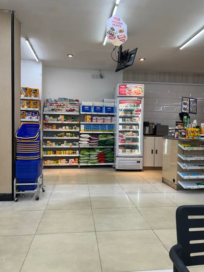The image shows the interior of a convenience store, likely in Depok, Indonesia, with aisles of products and a refrigerated display. In the foreground, a stack of blue shopping baskets is on a wheeled cart. To the left, shelves are stocked with baby formula and other packaged goods. A white refrigerated display case is centrally located, showcasing various food items, including what appears to be pre-packaged meals. Behind the display case, there is a coffee machine and a counter area with more shelves stocked with snacks and beverages. A sign hanging from the ceiling advertises "FROZEN BENTO." The floor is tiled and reflective. There are no people visible in the frame.