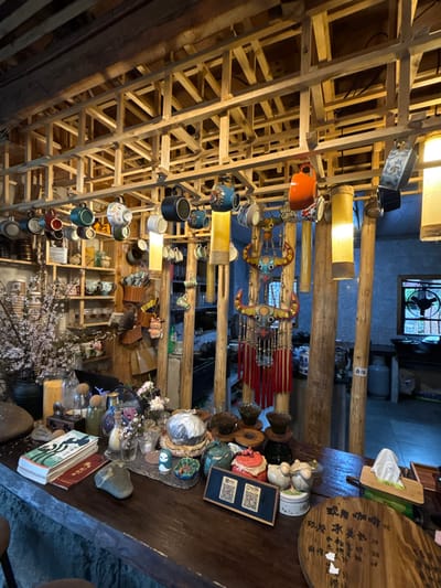 The image depicts the interior of a rustic establishment, likely a tea house or cafe, situated in Gongtan, China. The scene is characterized by extensive use of natural wood and an eclectic display of decorative and functional objects.

The ceiling features a dense lattice structure of light-colored wooden beams, from which numerous ceramic mugs of various designs and colors are suspended. Interspersed among the mugs are several cylindrical, translucent yellow fabric lanterns that emit a warm glow. Larger, darker wooden beams are visible above this lattice.

Structural elements include several robust, vertically oriented wooden posts, which appear to support the overhead structure. Between two central posts hangs a large, ornate, multi-tiered decorative piece resembling a stylized animal skull or mask, primarily red, blue, and yellow, with prominent red tassels descending from its lower section.

On the left, a tall vase containing blossoming branches with numerous small, light-colored flowers is positioned on a wooden surface. Behind it, a multi-shelf wooden unit displays an assortment of ceramic items, baskets, and small containers.

The foreground is dominated by a dark, polished wooden counter laden with various items. These include stacks of books, a smooth grey stone, glass jars containing seeds or grains, small vases with flowers, and a collection of ceramic tea ware and decorative figurines. A set of dark ceramic teacups or coffee drippers rests on individual wooden stands. A small wooden sign displaying two QR codes is prominently placed on the counter. To the right, a white tissue box is visible, alongside a circular wooden plaque with handwritten Chinese characters, likely a menu.

In the background, a window with a dark frame allows a glimpse of an external or adjacent space, revealing a metal grate or railing. Below the window, a light-colored gas cylinder is partially visible. No individuals are present in the scene.