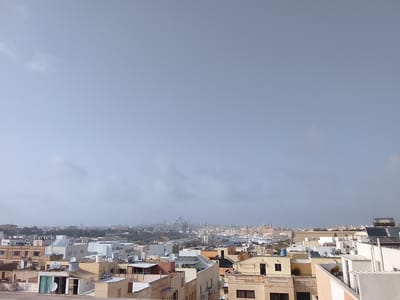 The image shows a cityscape of Fgura, Malta, under a cloudy sky. The foreground is dominated by rooftops of residential buildings, a mix of white and beige colors. Many of the buildings have satellite dishes and water tanks on their roofs. In the mid-ground, the city stretches towards the horizon, with industrial cranes visible in the distance. In the background, the skyline is hazy and slightly blurred due to the overcast conditions. No people or specific interactions are visible in the frame.