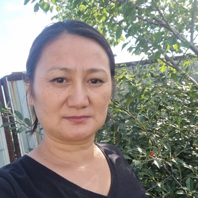 A person with dark hair and brown eyes is shown in a close-up, outdoor shot. They are wearing a black t-shirt. The background consists of green leaves and branches of a tree, with a glimpse of a light-colored sky and a metal structure that appears to be a fence or corrugated metal siding. There is a single red berry visible on a branch in the upper right portion of the frame. The lighting suggests it is daytime, possibly sunny, as there are bright highlights on the person's skin and some leaves. The person's facial expression is neutral. The overall scene suggests a casual, possibly domestic or natural setting.