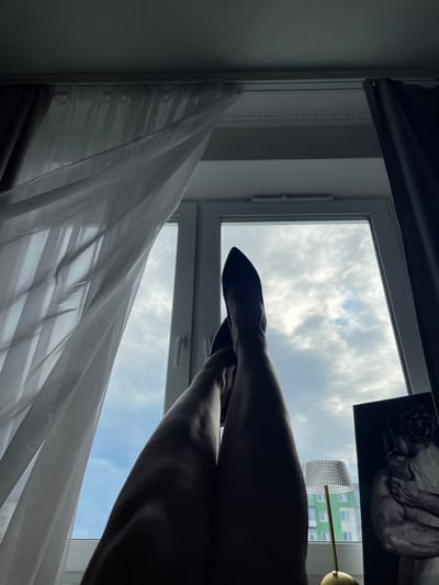 The image depicts a view from indoors, looking out of a window. Two silhouetted legs wearing black pointed-toe heels are stretched upwards, positioned in the foreground and obscuring a significant portion of the window. The window looks out onto a cloudy sky. Sheer white curtains are draped to the left, and dark curtains are to the right of the window. To the right of the window, a small lamp with a textured glass shade and a gold base is visible, as well as a framed artwork of two embracing figures. In the background, through the window, a green multi-story building is visible. The scene appears to be an interior space in a residential setting, possibly an apartment, located in Novogorelovo, Russia, given the presence of a typical Russian apartment building in the background.