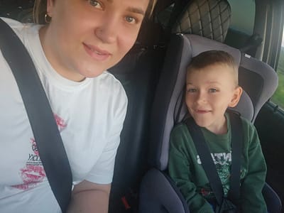 The image shows a selfie taken inside a car. A woman with blonde hair and blue eyes is in the foreground, wearing a white t-shirt with red lip prints and text. She has a nose ring and hoop earrings. Next to her, a young boy with blonde hair and blue eyes is sitting in a car seat. He is wearing a green sweatshirt with a graphic design. The car interior is visible, including the car seat and headrest, and a seatbelt crosses the woman's chest. The background outside the car appears to be a landscape, possibly rural, with a hint of green foliage and a bright sky, suggesting it is daytime. The image composition and the candid expressions of the subjects suggest a casual moment, likely during travel. There is no indication of the location being the city of Dubrova, Russia, within the image itself.