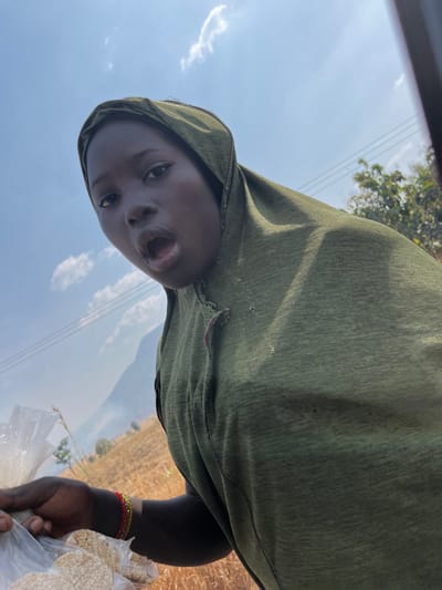 An outdoor image taken in Nandu, Nigeria, features a dark-skinned individual looking directly into the camera with their mouth open. The person wears an olive-green hooded garment, which shows small tears or fraying near the chest. Their visible right arm holds several transparent plastic bags containing light-colored, circular food items. Thin, multi-colored bracelets (red, yellow, green) are visible on their right wrist. The background consists of a bright blue sky with scattered white clouds, a dry, yellowish-brown field, and distant hazy hills or mountains. Overhead utility wires are visible in the middle distance. A dark vertical element, likely part of a vehicle frame, is present on the far right side of the image.