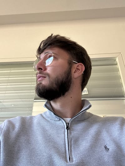 The image depicts an indoor scene. While the user specified the location as Lièpvre, France, there are no visual cues within the image to corroborate this information.

A male individual is positioned in the foreground, captured in a mid-shot from a low angle. He has short, dark brown hair and a full, dark brown beard. He wears round, thin-framed metallic glasses and a white earbud in his right ear. His head is turned slightly to the left, and his gaze is directed upwards and to the left. He is wearing a light grey quarter-zip sweater with a textured knit pattern. A small, dark embroidered logo, consistent with the Ralph Lauren polo player emblem, is visible on the left chest area of the sweater.

The background consists of a light-colored wall on the upper left and a white-framed window on the right. Horizontal white Venetian blinds are visible through the window. Faint, dark lettering spelling "AFF" is visible on the windowpane in the lower right portion of the background. Lighting appears bright and even, likely originating from the window.