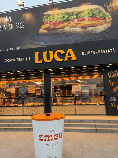An exterior, ground-level photograph depicts the facade of a "LUCA" establishment in Agigea, Romania. Above the storefront, a large black billboard features a prominent image of a sandwich with breaded cutlets, melted cheese, lettuce, and tomatoes, accompanied by Romanian text including "FACEM BUN, BUN SE FACE" and "SANDVIȘ CU ȘNIȚEL". Water droplets are visible on the billboard's surface. Below the billboard, the store's primary signage displays "LUCA" in large orange letters, flanked by "TRADIȚIE" and "REINTERPRETATĂ" in white text on a black background. The storefront consists of multiple glass panels, revealing interior display cases filled with various baked goods and pastries. Internal signage displaying "ZMEU" branding and additional Romanian text is visible through the glass. In the immediate foreground, centered in the lower frame, a white cylindrical stand with an orange lid features an integrated ashtray and bears the orange text "CAFEA ZMEU ARTIZANALĂ". The ground consists of light-colored square paving tiles, leading to a series of steps ascending to the store entrance. The sky above is overcast. No individuals are visible.