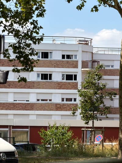 The image depicts a residential building in Évry-Courcouronnes, France, under a partly cloudy sky. The building is constructed with brick and white sections, featuring multiple floors with rows of windows. Some windows have white shutters that are closed, while others are open, revealing dark interiors. A balcony with a railing extends from one section of the building.
In the foreground, foliage from trees partially obscures the view. To the left, the front of a white vehicle is visible, with a VW logo on its grille. To the right of the building, another tree stands, and in front of it, a circular red and blue sign with a diagonal cross is mounted on a wooden frame, indicating a parking prohibition. The ground is covered with grass and dry vegetation. The overall scene suggests a typical urban residential environment.
