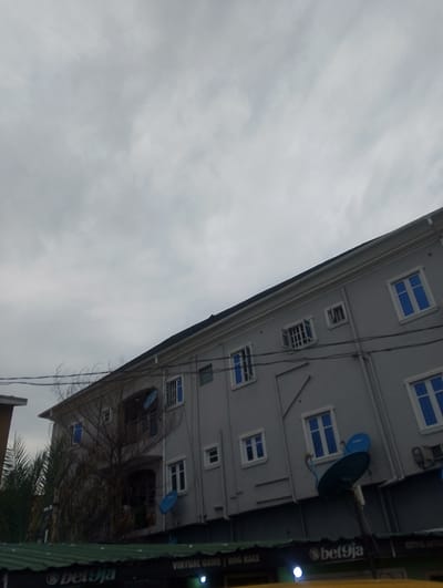 The image shows a multi-story building with a grey facade under a cloudy sky. The building has numerous windows, some with blue tint. There are satellite dishes attached to the exterior of the building. In the foreground, a green awning is visible with text that includes "bet9ja" and "VIRTUAL GAME DOG RACE". The overall scene depicts an urban environment, possibly a commercial or residential building in Alagbede, Nigeria. There are no people visible in the image. The weather appears overcast.