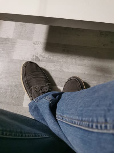 The image shows a close-up, overhead view of a person's legs and feet wearing dark gray canvas boat shoes and blue denim jeans. The shoes have a light-colored sole and dark laces. The jeans are folded and draped across the legs. The person is sitting on a floor with a light-colored wood-grain pattern. A white surface, possibly a table or shelf, is visible in the background, casting a shadow on the floor. There is no discernible location in the city of Bourg-lès-Valence, France, within the image.