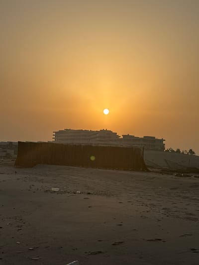 A wide shot captures a beachfront scene in Ajman, United Arab Emirates, during either sunrise or sunset, indicated by the low, bright orange sun positioned in the hazy sky. The sun is partially obscured by atmospheric conditions, giving the sky a muted, yellowish-orange gradient. In the mid-ground, a long, textured barrier constructed from what appears to be natural reeds or straw runs horizontally across the frame. Behind this barrier, the silhouettes of several unfinished or abandoned multi-story buildings with flat roofs and exposed concrete structures are visible. The buildings suggest a construction site or a development project that has been halted. The foreground consists of a sandy beach, with footprints and scattered debris, including plastic bottles, visible on the sand. The overall impression is one of an undeveloped or neglected coastal area.