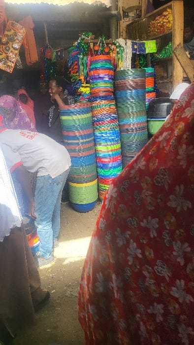 The image displays an active market stall in Fagge D1, Nigeria, beneath a corrugated metal roof structure. In the immediate foreground, the right side is occupied by a partial view of a person wearing a red garment with a white floral pattern. To the mid-left, a male subject wearing a white short-sleeved t-shirt with red trim and light blue jeans bends forward, facing away from the camera. Partial text "NEW HEA" is visible on his shirt. To his left, another male subject with dark skin, dressed in a dark top, smiles broadly towards the left, with his right hand raised near his face. In the background-left, two individuals wearing purple and pink head coverings are partially visible. Multiple tall, vertical stacks of assorted plastic basins or baskets are prominent in the central and right background, displaying various colors including blue, green, yellow, orange, red, and brown. Above, various textiles, bags, and green decorative string lights hang. Shelves behind the stacks hold additional merchandise. A black plastic container and a green plastic container rest on the floor to the mid-right. The ground is an uneven, light-colored surface.