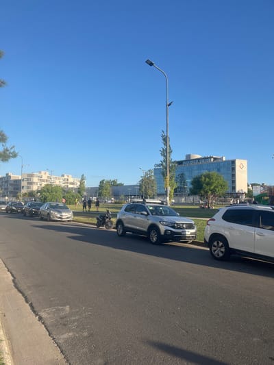 The image depicts an urban outdoor scene in Partido de Quilmes, Argentina, under a clear blue sky, suggesting daytime with long shadows indicating late afternoon. In the foreground, an asphalt road runs horizontally, bordered by a concrete curb on the left. Approximately six vehicles are parked in a line on the right side of the road. The most prominent vehicle is a silver Volkswagen T-Cross SUV with a visible Argentinian license plate "AE 531 FP" and roof rails. A dark-colored scooter or motorcycle is parked in front of the T-Cross. Other parked vehicles include a partially visible white SUV and a dark grey sedan.
A wide, grassy median or park area separates the road from background structures. A tall, grey metal streetlamp with an LED light fixture stands prominently within this grassy area, casting a shadow diagonally across the grass. Approximately 10-15 individuals are distributed across the grassy expanse. Some are walking or standing, while others are gathered in small groups, with several appearing to be seated or engaged in a recreational activity in the mid-distance. Trees with green foliage are scattered throughout the park and near the buildings.
In the background, a modern, multi-story building with a reflective glass facade and a distinctive reddish-brown diamond logo (partially obscured, possibly "OMNIUM" or "OHI") is visible on the right. To its left, another multi-story, lighter-colored building with multiple windows or balconies stands. Further structures are visible in the distant background. No active vehicle movement is observed, and people's actions are consistent with casual park leisure.