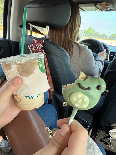 A person is driving a car in Santa María Moyotzingo, Mexico, and another person in the passenger seat is holding a Starbucks cup with a green straw in their left hand and a partially eaten frog-shaped cake pop in their right hand. A brown wallet or purse is visible near the Starbucks cup. The driver has long brown hair and is wearing a gray sweater. The passenger is wearing blue jeans. Part of the landscape is visible through the front window. A scarf is on the passenger seat behind the Starbucks cup.