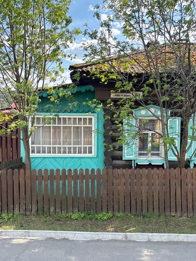 The image depicts a wooden house in Овсянка, Russia. The house is painted in a blue-green color and has white framed windows with lace curtains. A dark brown wooden fence runs along the side of the house, bordering a street. A tree with sparse green leaves partially obscures the view of the house. A sign that reads "УЛ. НАБЕРЕЖНАЯ" is also visible on the house. The sky is visible through the tree branches, with scattered clouds. The street is made of asphalt.
