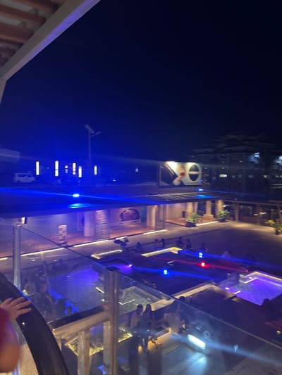 The image depicts a nighttime scene in what appears to be a modern establishment, possibly a shopping mall or entertainment complex, in Davao City, Philippines. The perspective is from a slightly elevated position, looking down and across a multi-level structure.

The primary subjects are the architectural elements and lighting. Dominant features include glass railings, floors, and ceilings, reflecting the vibrant blue and red lights that illuminate the space. Staircases and escalators are visible, suggesting movement and accessibility within the building. In the background, a tall building with illuminated windows indicates an urban setting.

There are hints of people, seen as blurred figures or reflections, particularly in the lower right portion of the image where a hand is visible resting on a railing. The lighting creates a dynamic and somewhat futuristic atmosphere. The time of day is clearly night, evidenced by the dark sky and the reliance on artificial lighting.

Notable details include the use of neon-like blue and red lights, which create a striking visual contrast. Planters with greenery are interspersed throughout the different levels, adding a touch of nature to the otherwise sleek, modern design. A large circular sign with some lettering is visible on one of the buildings, though the text is not legible. The overall impression is one of a lively and contemporary public space.
