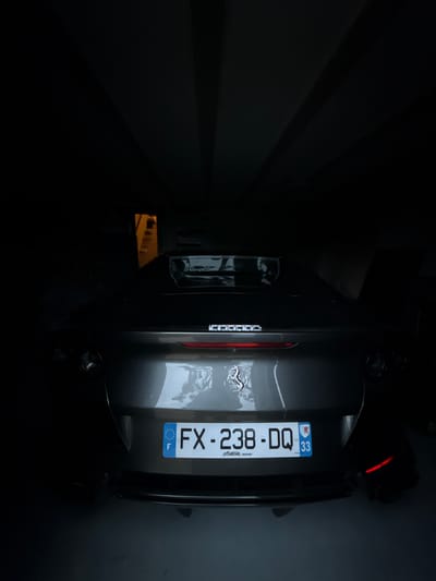 The image displays the rear section of a dark grey Ferrari sports car, specifically identifiable by the Prancing Horse emblem, parked within a dimly lit indoor space. The vehicle is centrally aligned, presenting its tail lights, quad exhaust tips, and a prominent rear diffuser. A French license plate, "FX-238-DQ," is affixed to the trunk lid; it features the national identifier "F" on the left and the departmental code "33" for Gironde, accompanied by the Nouvelle-Aquitaine coat of arms, on the right. Below the license plate, the text "matin .SENS" is present. The environment is a dark garage or parking structure, characterized by overhead architectural elements and minimal ambient light. A distant, warmer light source is discernible through an opening to the left of the vehicle. The floor is a dark, mildly reflective surface. This scene is situated in a private garage in Lège-Cap-Ferret, France, consistent with the license plate's regional identification. No persons are visible, and the vehicle is stationary.