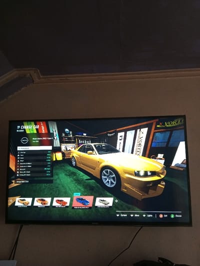 The image displays a television screen showing a car customization menu in a racing game. The primary focus is a golden-yellow Nissan Skyline R34 with a wide body kit, showcased in a virtual garage. The "Change Car" menu lists parameters on the left, while various car models for selection are displayed along the bottom. The game interface includes options such as camera, move, lights, exit, and choose.