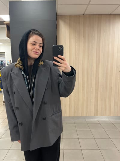 The image depicts a woman taking a mirror selfie inside a commercial establishment in Urvillers, France. The subject, a female with light brown hair and a subtle smile, occupies the central frame, looking directly into the smartphone held in her left hand. She is wearing an oversized dark grey double-breasted blazer with black buttons, a dark knitted hood with a leopard print trim, and a zippered dark top underneath. Her fingernails are painted a light color with a subtle pattern. The indoor environment features a dark vertical panel on the left, a light wood-paneled wall on the right, a white square-tiled ceiling, and a light grey square-tiled floor. Distant reflections of what appear to be clothing racks or store fixtures are faintly visible in the background.