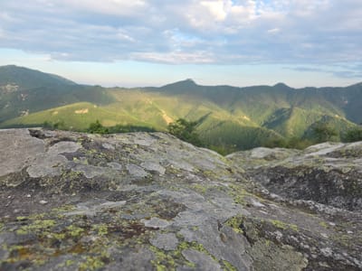 The image depicts a panoramic view of a mountainous landscape, likely taken from a high vantage point in the Vitosha mountain range, near Sini Vrah, Bulgaria. In the foreground, a large, textured rock covered in lichen dominates the lower portion of the frame. The rock exhibits patches of green and gray lichen, showcasing its weathered surface. Beyond the rock, rolling hills covered in lush green vegetation extend into the distance. The hills are densely forested and appear to be part of a rugged mountain range. Sunlight illuminates some of the slopes, creating a contrast between light and shadow. In the far background, several peaks of the mountain range are visible, silhouetted against a pale blue sky with scattered clouds. The sky is partly cloudy, with soft white clouds drifting across the horizon. The overall atmosphere is serene and natural, suggesting a clear day with good visibility in the mountainous terrain.