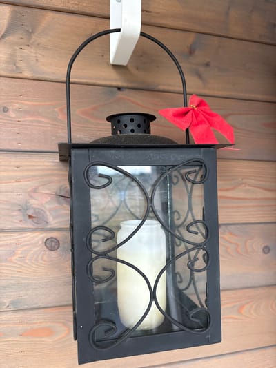 An outdoor lantern hangs on a wooden wall. The lantern is black and rectangular with glass panels and ornate metal scrollwork. A white candle is visible inside the lantern. A small red bow with a gold bead in the center is tied to the top handle of the lantern. The wooden wall is made of horizontal planks with visible grain and knots. The scene is outdoors.