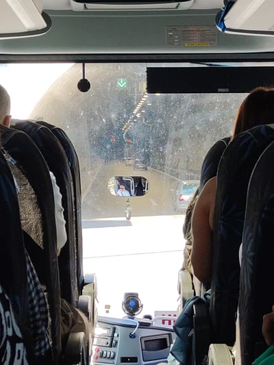 The image is taken from inside a bus, looking forward through the windshield and a side window. The bus is driving into a tunnel. The tunnel appears to be well-lit with overhead lights and traffic lights visible in the distance. There are cars in front of the bus, also heading into the tunnel. A rearview mirror inside the bus shows a man wearing sunglasses and a white shirt, presumably the bus driver. Seats of the bus are visible on the left and right sides of the image, some occupied by passengers. The exterior of the bus, including a dashboard with a small fan, is also visible. The Municipal Unit of Makrichori, Greece, is known for its scenic routes and tunnels, suggesting this could be a view within that region.