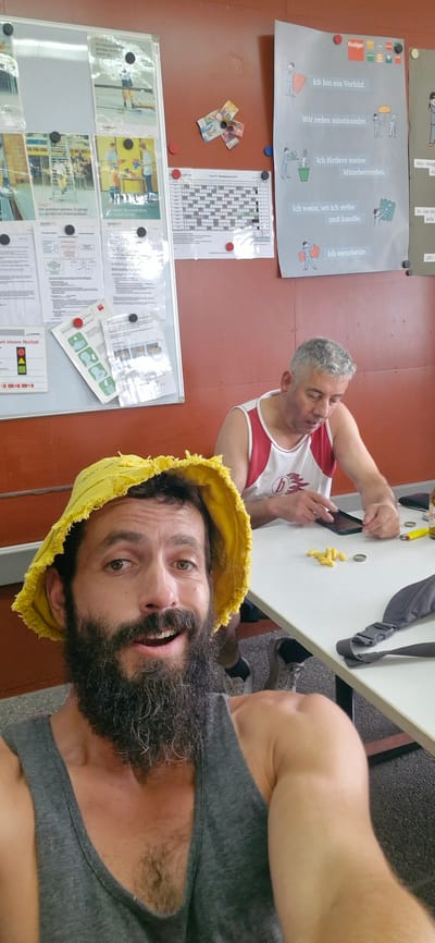 In Uetendorf, Switzerland, two men are seated at a white table. The man in the foreground, with a prominent beard and a yellow bucket hat, takes a selfie. He wears a grey tank top. The man in the background, with grey hair, sports a white tank top with red trim. He appears to be using a smartphone. On the table, between the two men, are scattered yellow corn snacks, a mobile phone, a yellow lighter, and a bottle cap. The environment includes a bulletin board covered with various papers and posters on the left and a large poster with German text and illustrations on the right. The wall behind the men is a warm, reddish-brown color. The floor is carpeted with a dark, speckled pattern.