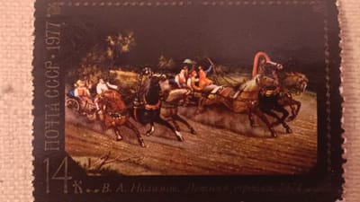 The image is a postage stamp featuring a painting of a troika, a Russian horse-drawn sleigh or carriage, with three horses abreast. The troika is depicted in motion, with the horses' legs blurred to suggest speed. Several figures, dressed in traditional attire, are seated in the troika and on a nearby carriage, some holding reins or whips. The scene appears to be set in a rural or natural environment, with trees and a dirt track visible. The stamp itself is dark in color, with white lettering on the left side that reads "ПОЧТА СССР 1977" (POST USSR 1977) and a number "14". Below the painting, there is more Cyrillic text, partially obscured, that includes "...В. А. Налимов. Летняя тройка 1974" (...V. A. Nalimov. Summer Troika 1974). The stamp has a perforated edge. There is no information within the image or its text that suggests a location in Onaldy, Turkmenistan.
