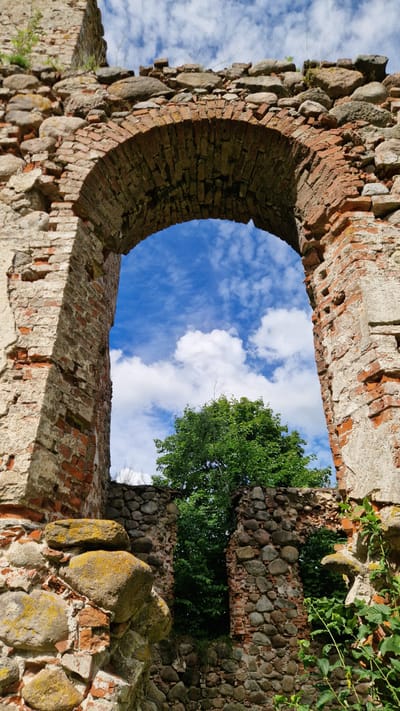 The image shows a view through an ancient brick archway of ruins located in Embūte, Latvia. The archway, constructed from red bricks, is partially crumbled, revealing rough stones and mortar. Through the arch, a vibrant green tree is visible against a bright blue sky with scattered white clouds. The ruins are overgrown with greenery, suggesting abandonment and the passage of time. The overall scene evokes a sense of history and nature reclaiming man-made structures.