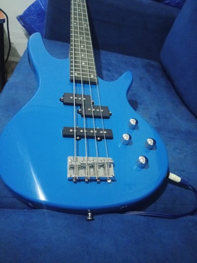 An electric bass guitar with a bright, glossy blue finish is positioned diagonally in the frame, resting on a textured blue upholstered surface, likely a sofa or couch. The instrument features a black fretboard with white fret markers and a headstock that extends out of view at the top. It is a 4-string bass, with four metal strings running over a chrome bridge and extending past two black pickups. The pickups appear to be a P/J configuration, with a split-coil (P-style) pickup in the mid-body position and a single-coil (J-style) pickup closer to the bridge.
Four chrome control knobs are visible on the lower bout of the guitar's body. An instrument cable, with a white-tipped connector, is plugged into the output jack located on the bottom edge of the bass. The background is softly blurred, showing more of the blue upholstered furniture, suggesting an indoor setting. The lighting is even, highlighting the reflective surface of the guitar's body. There are no people or visible text in the image.