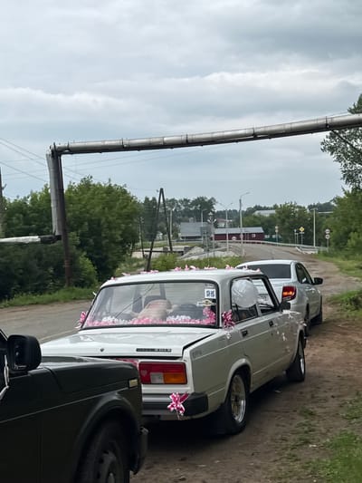 In Зюкайка, Russia, on an overcast day, a procession of cars, decorated with pink and white ribbons and flowers, drives along a dirt road. The lead vehicle, a dark SUV, is partially visible on the left. Behind it, a white sedan, a Lada 2105, is adorned with pink ribbons and flower garlands. A light silver sedan follows behind the Lada. The road is flanked by green trees and bushes. In the background, there is a large industrial pipe supported by a metal structure, power lines, and buildings. Traffic signs are visible in the distance.
